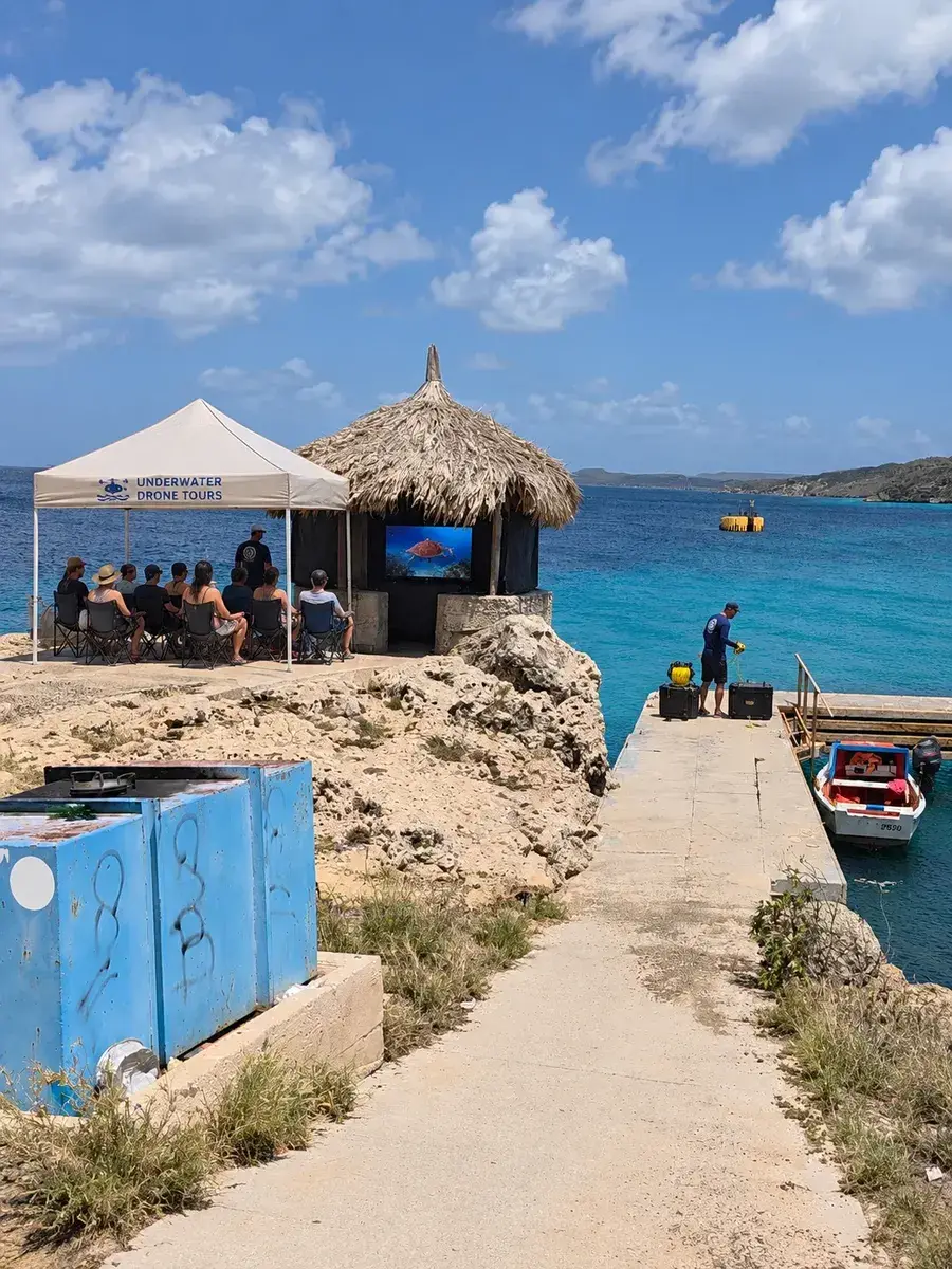 Guests seated under a canopy beside the palapa hut watching the live underwater feed on the big screen while the operator works at the pier